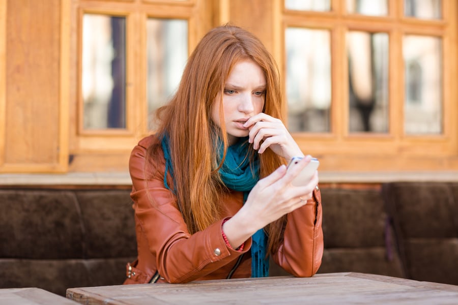 Embarrassed sad shoked young redhead lady reading message in mobile phone sitting in open air cafe