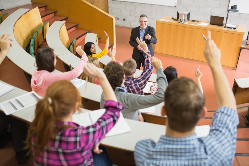 Rear view of students raising hands with a teacher in the college lecture hall
